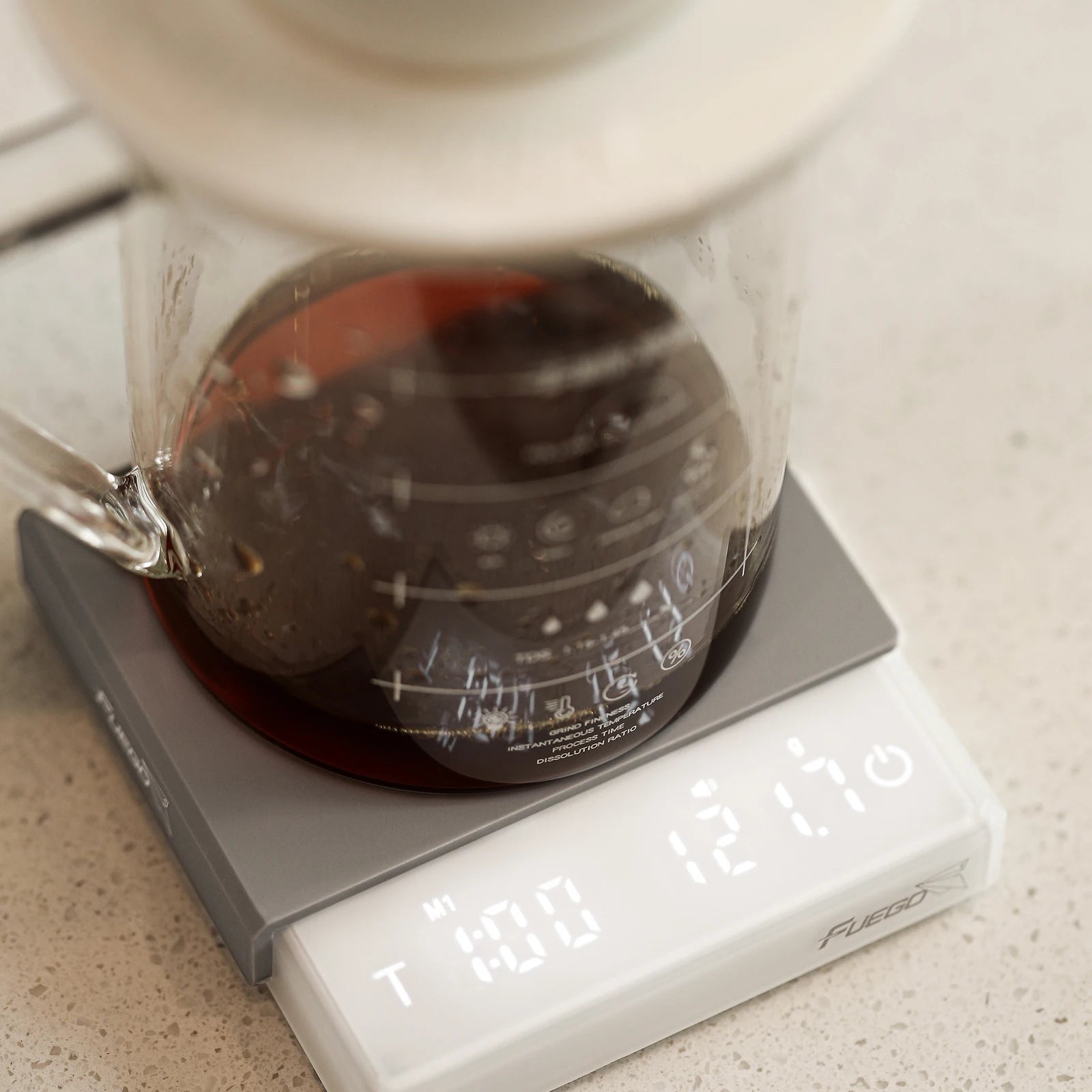 Glass coffee maker on a digital scale with coffee inside, on a light-colored surface.