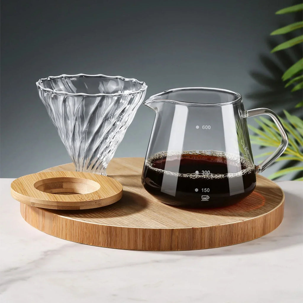 Glass coffee maker with coffee on a wooden tray, with a blurred green plant in the background.