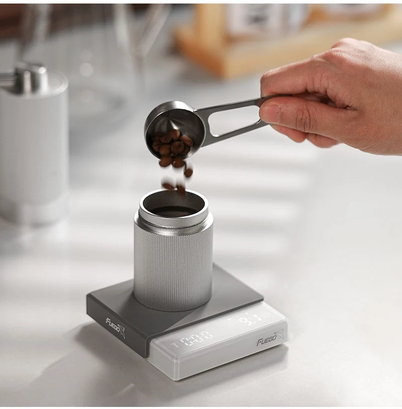 Person measuring coffee beans with a scoop over a coffee grinder on a digital scale.