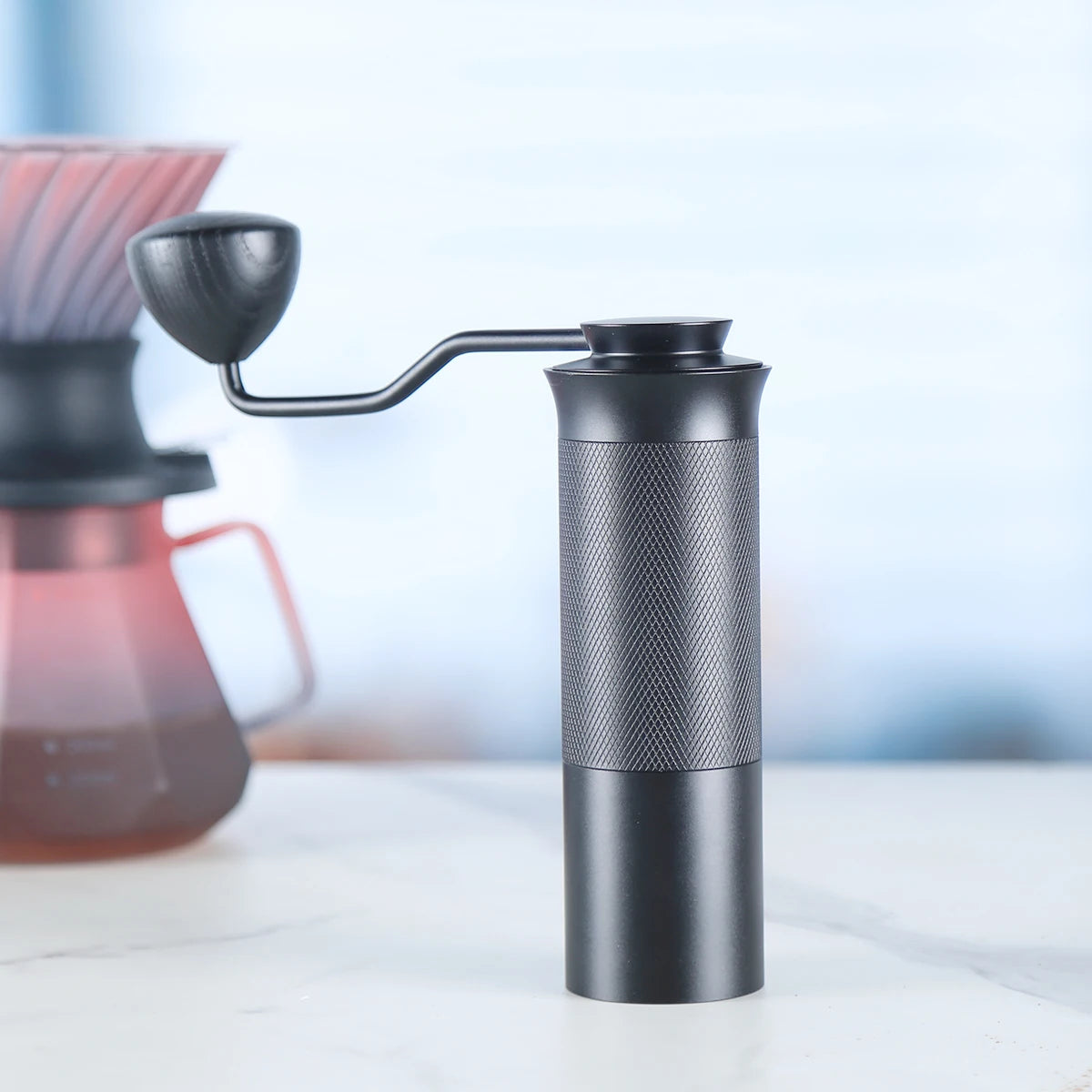 Handheld coffee grinder on a blurred background with a coffee filter and pot.