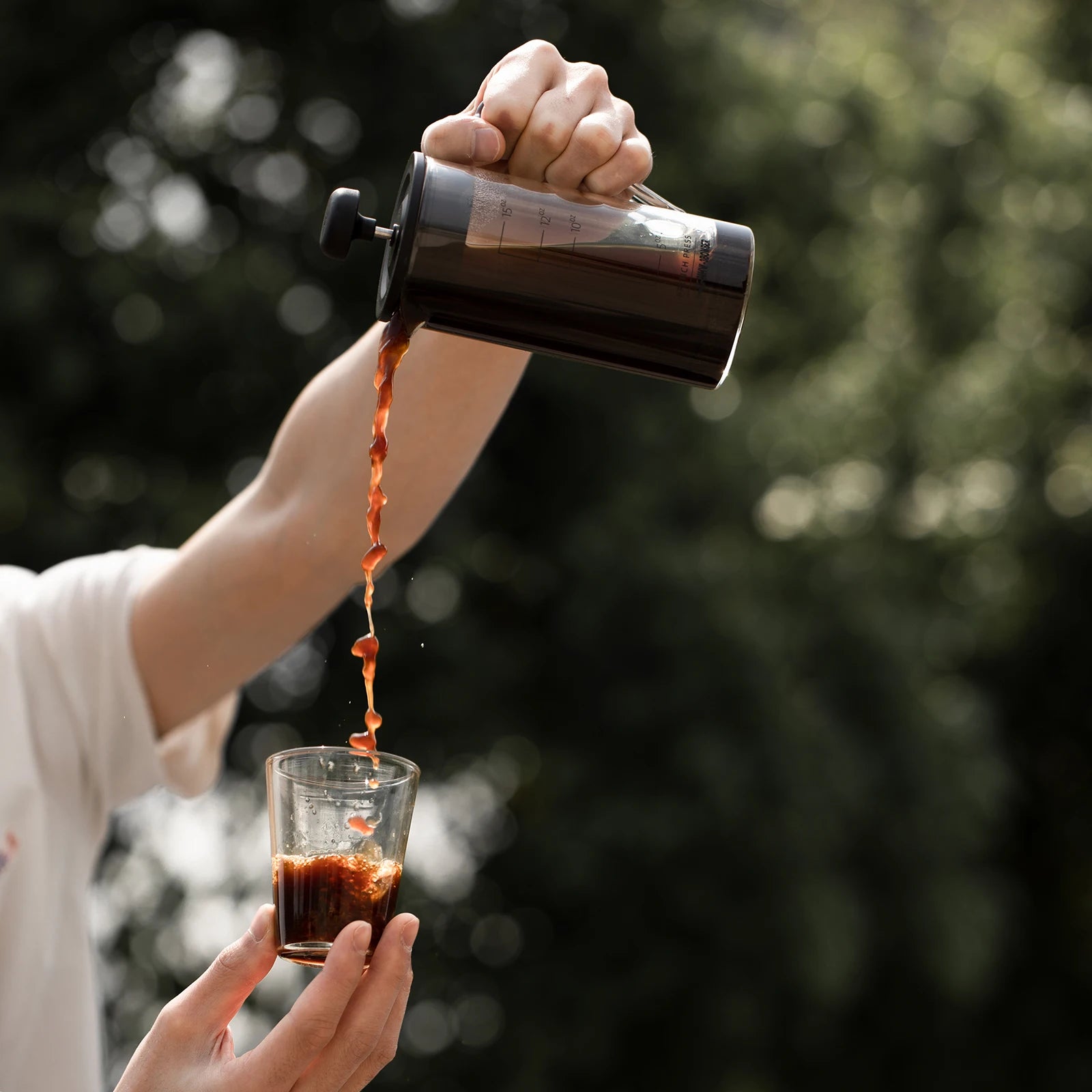 Person pouring coffee from a French press into a glass against a blurred natural background