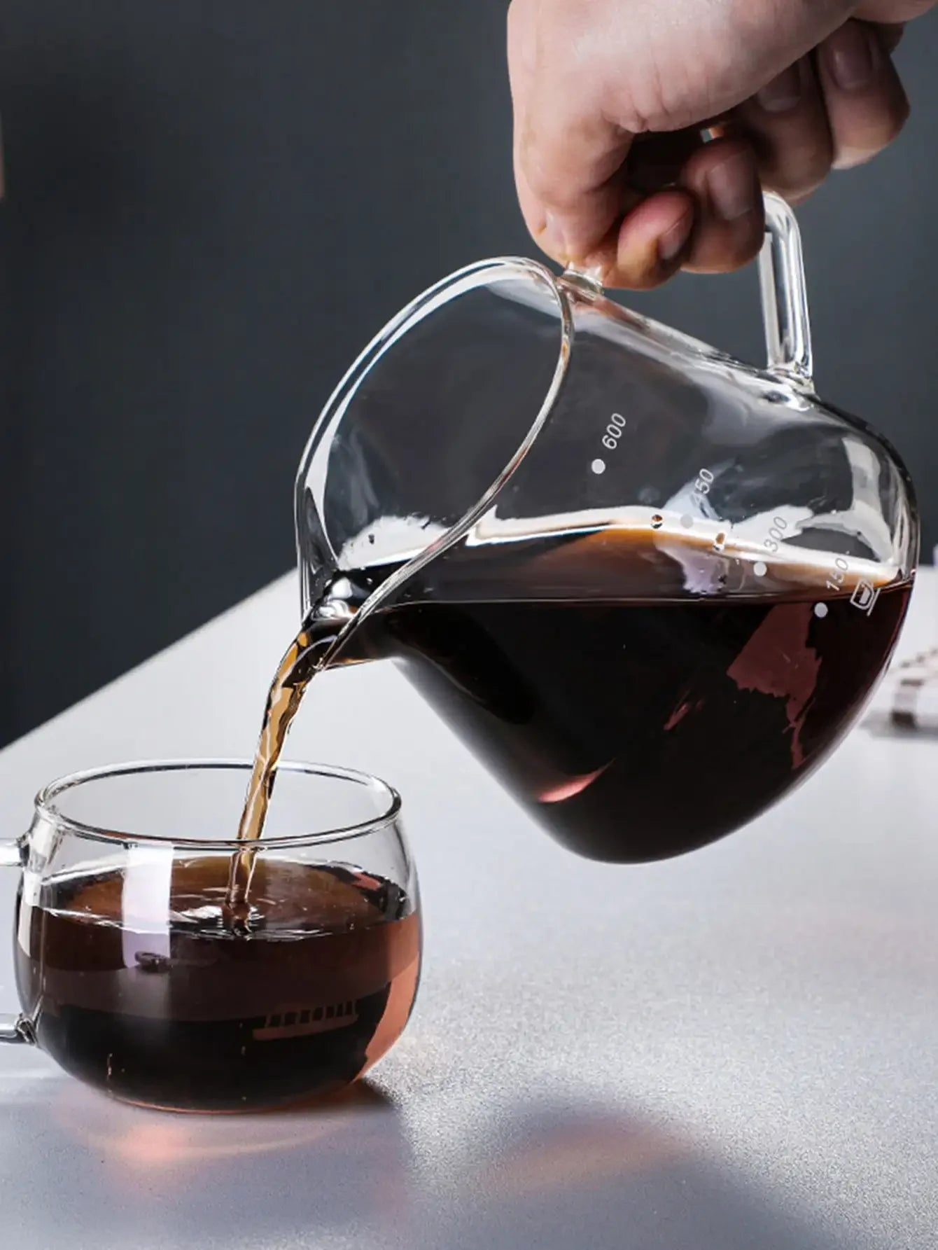 Person pouring dark liquid from a glass pitcher into a glass on a reflective surface with a dark background.