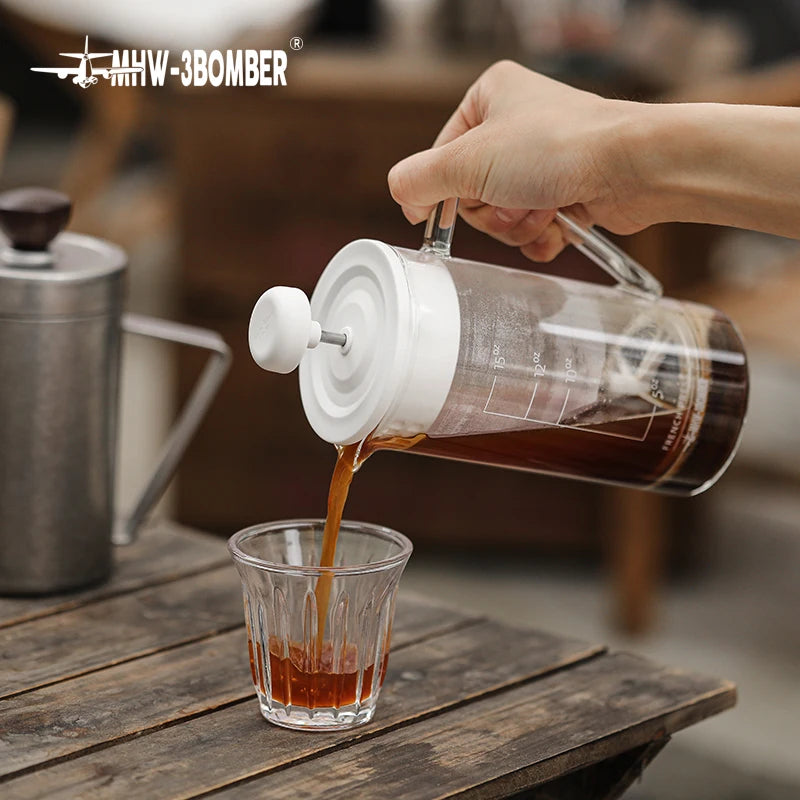 Person pouring coffee from a glass French press into a small glass on a wooden table.